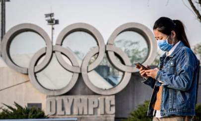 A woman wearing a mask walks before an Olympic rings sculpture at the national “Birds Nest” stadium in Beijing on March 23, 2020. (Nicolas Asfouri/AFP via Getty Images)